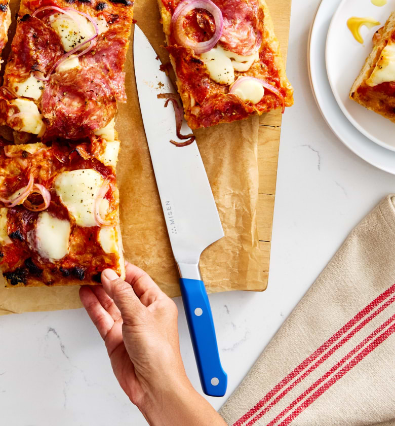 Misen chef's knife with blue handle cutting homemade Detroit-style pizza on wooden board