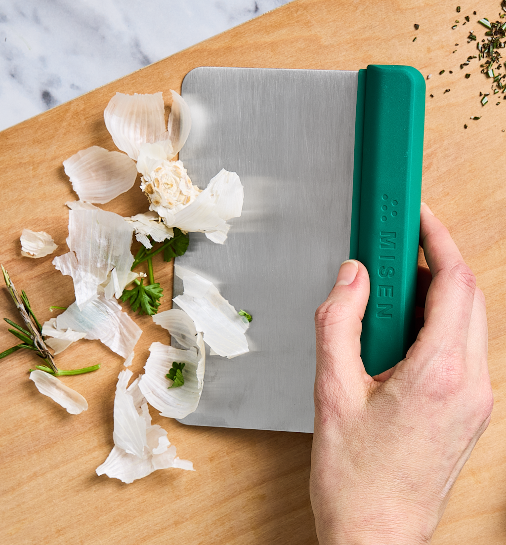 Misen bench scraper with green handle clearing garlic peels from wooden cutting board