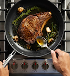 Basting a bone-in ribeye steak with butter, rosemary, and garlic in a skillet