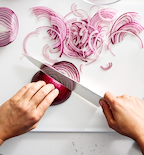 Hands slicing red onion into thin rings on white cutting board with chef's knife
