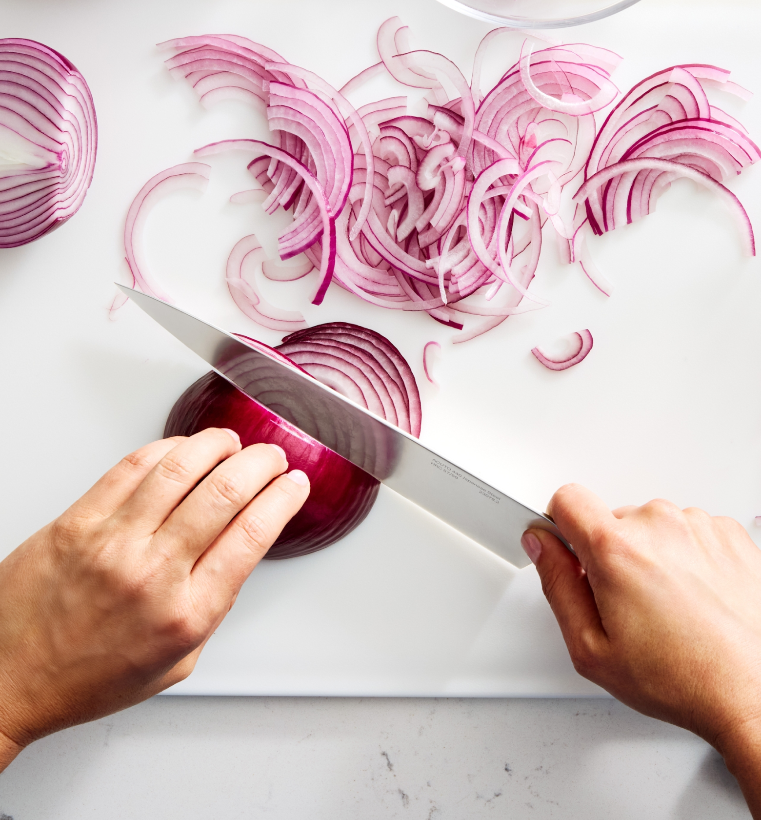 Hands slicing red onion into thin rings on white cutting board with chef's knife