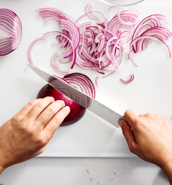 Hands slicing red onion into thin rings on white cutting board with chef's knife