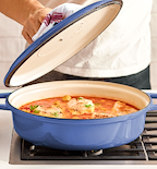 Chef lifting lid off blue enameled cast iron braiser with fish in tomato sauce on gas stovetop