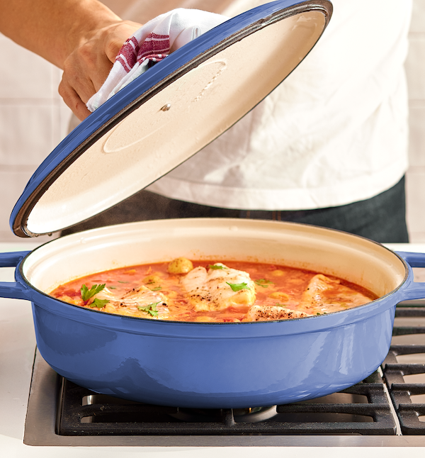 Chef lifting lid off blue enameled cast iron braiser with fish in tomato sauce on gas stovetop