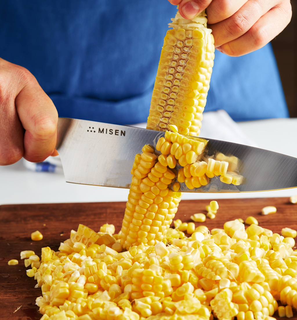 Cutting fresh corn kernels off the cob with a Misen chef's knife on a wooden cutting board