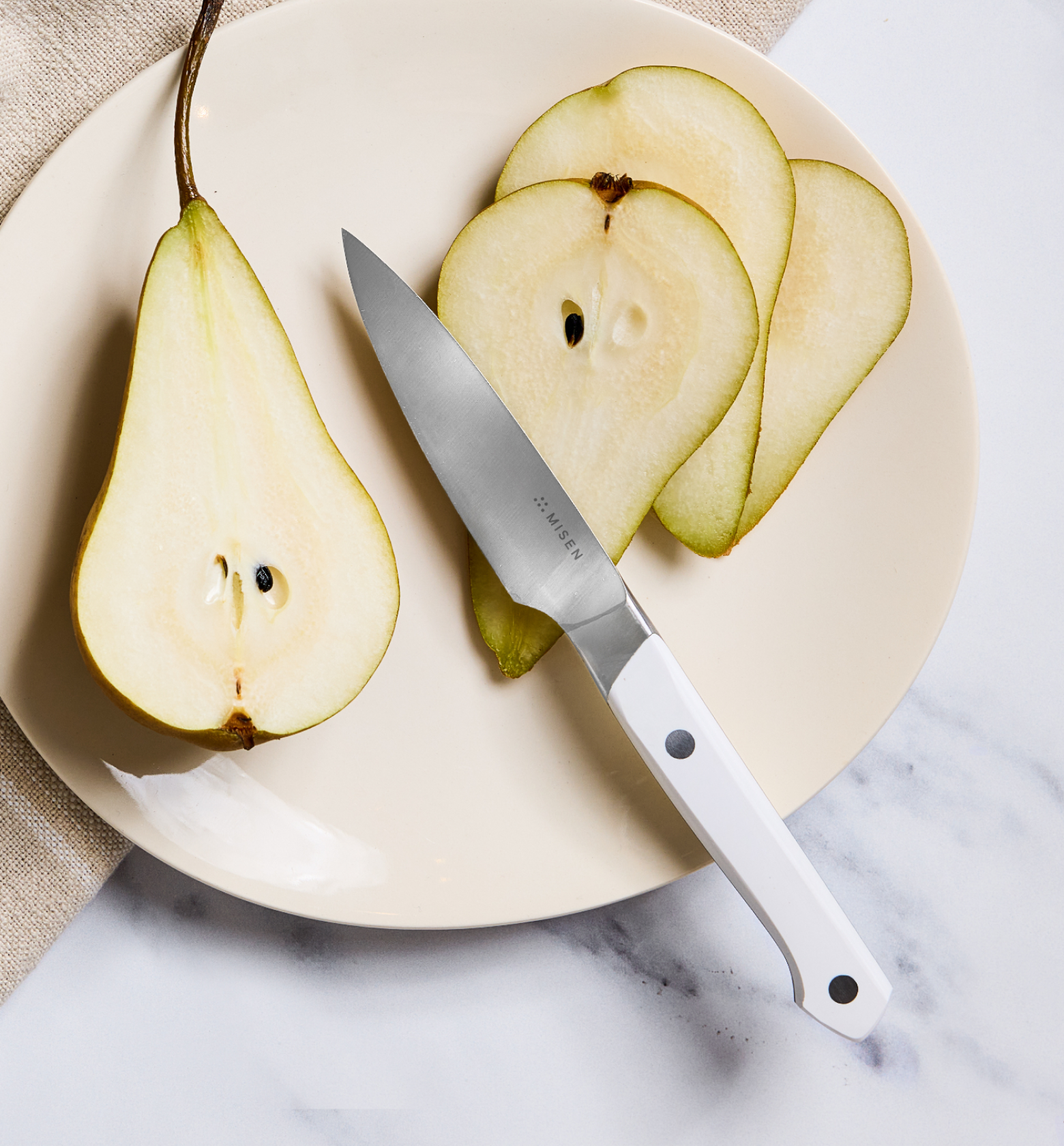 Misen paring knife with white handle beside sliced green pear on cream plate