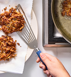 Crispy potato latkes being transferred from frying pan to paper towel-lined plate with slotted spatula