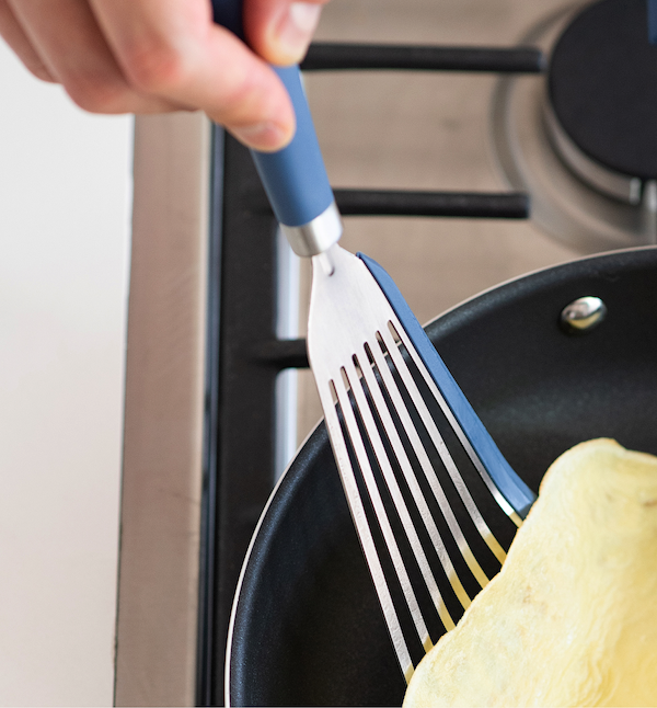 Blue-handled slotted spatula flipping an omelette in a nonstick frying pan on a gas stove