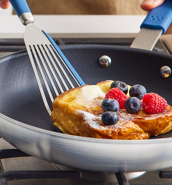 French toast topped with blueberries, raspberries, and butter cooking in a nonstick skillet with blue-handled spatula