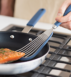 Fish spatula flipping seasoned salmon fillet in blue nonstick skillet on gas stovetop