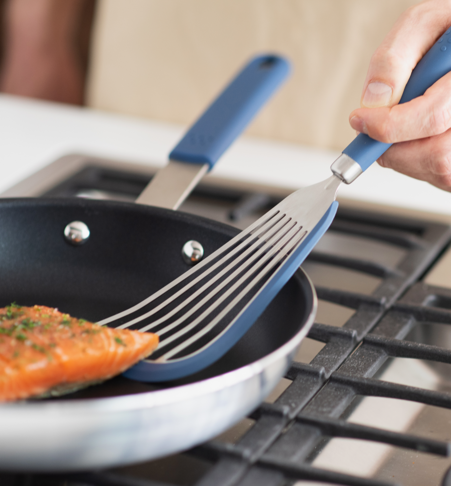 Fish spatula flipping seasoned salmon fillet in blue nonstick skillet on gas stovetop