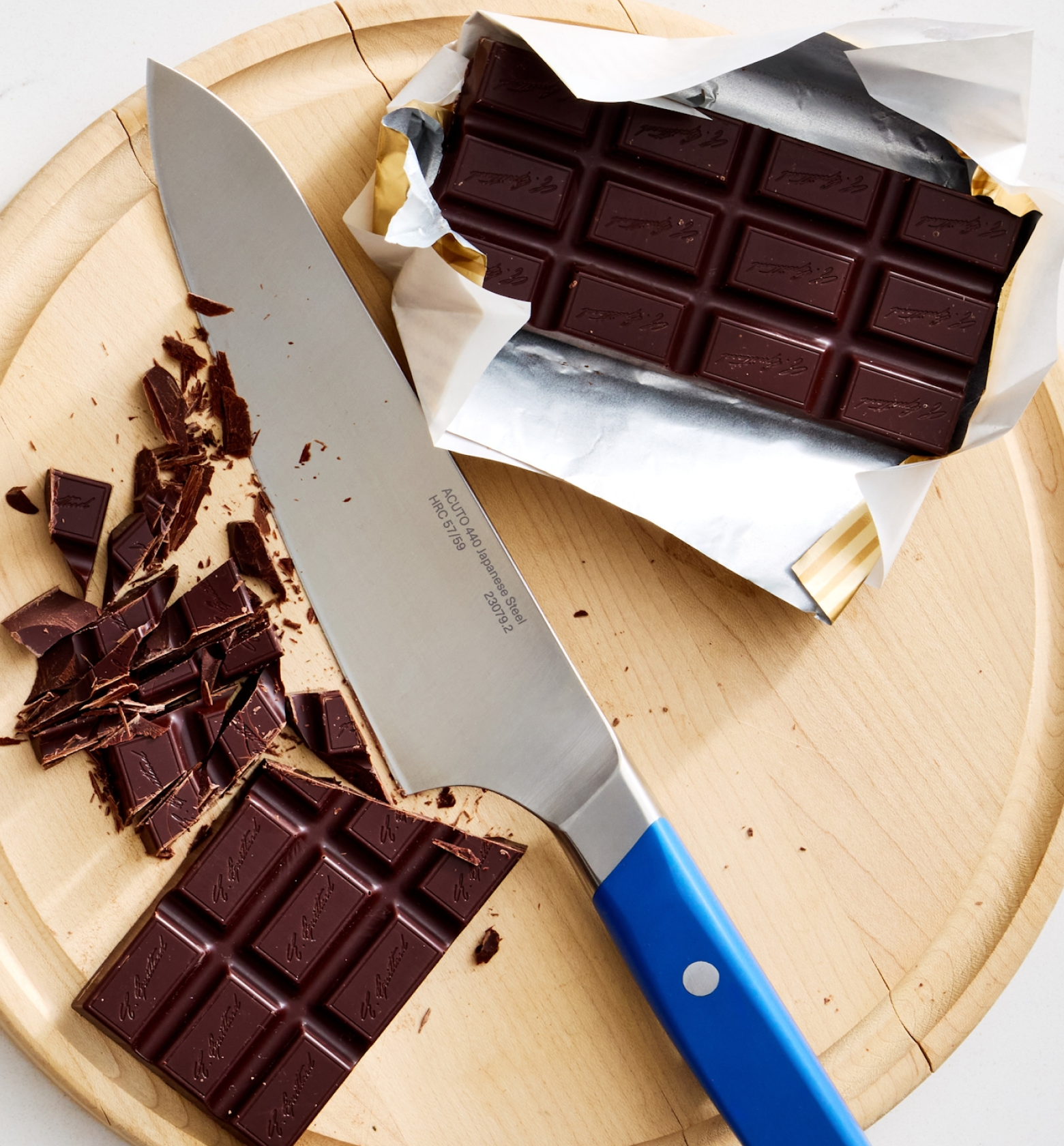 Dark chocolate bars being chopped with an Acuto 440 Japanese steel chef knife with blue handle on wooden cutting board