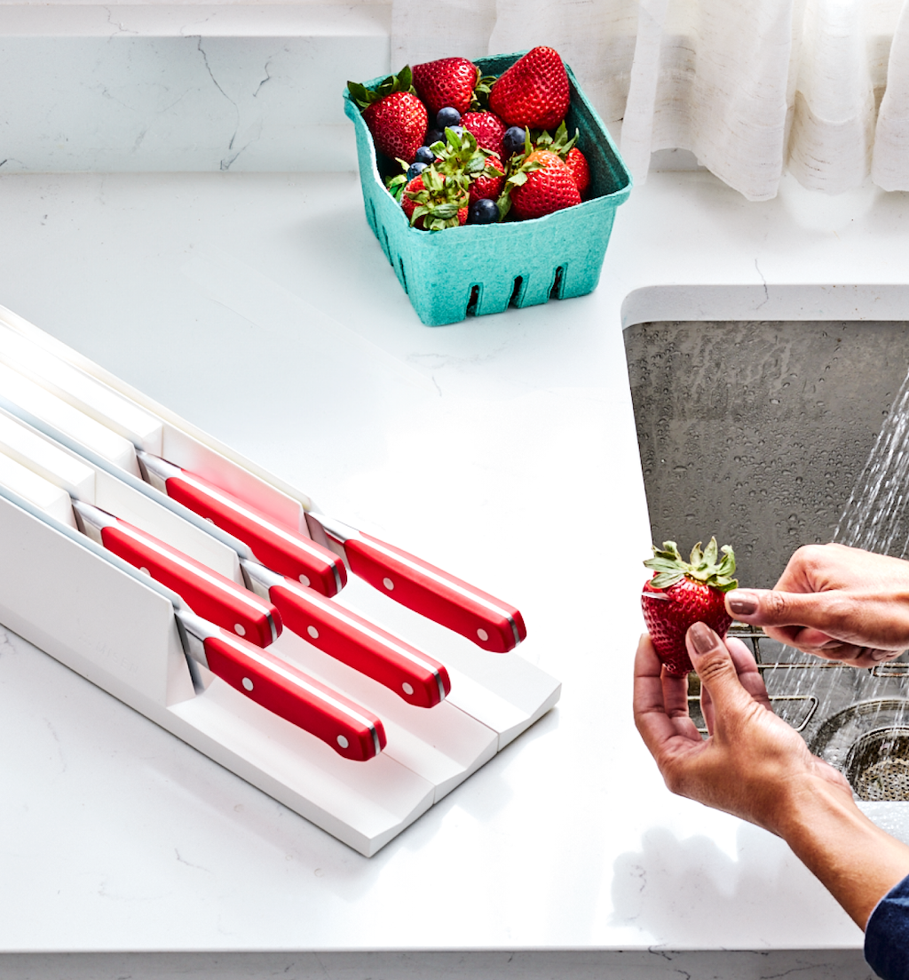 Red-handled kitchen knives in white knife block with fresh strawberries being rinsed at sink