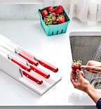 Red-handled kitchen knives in white knife block with fresh strawberries being rinsed at sink