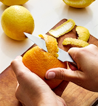 Hands peeling an orange with a paring knife on a wooden cutting board with lemon peels