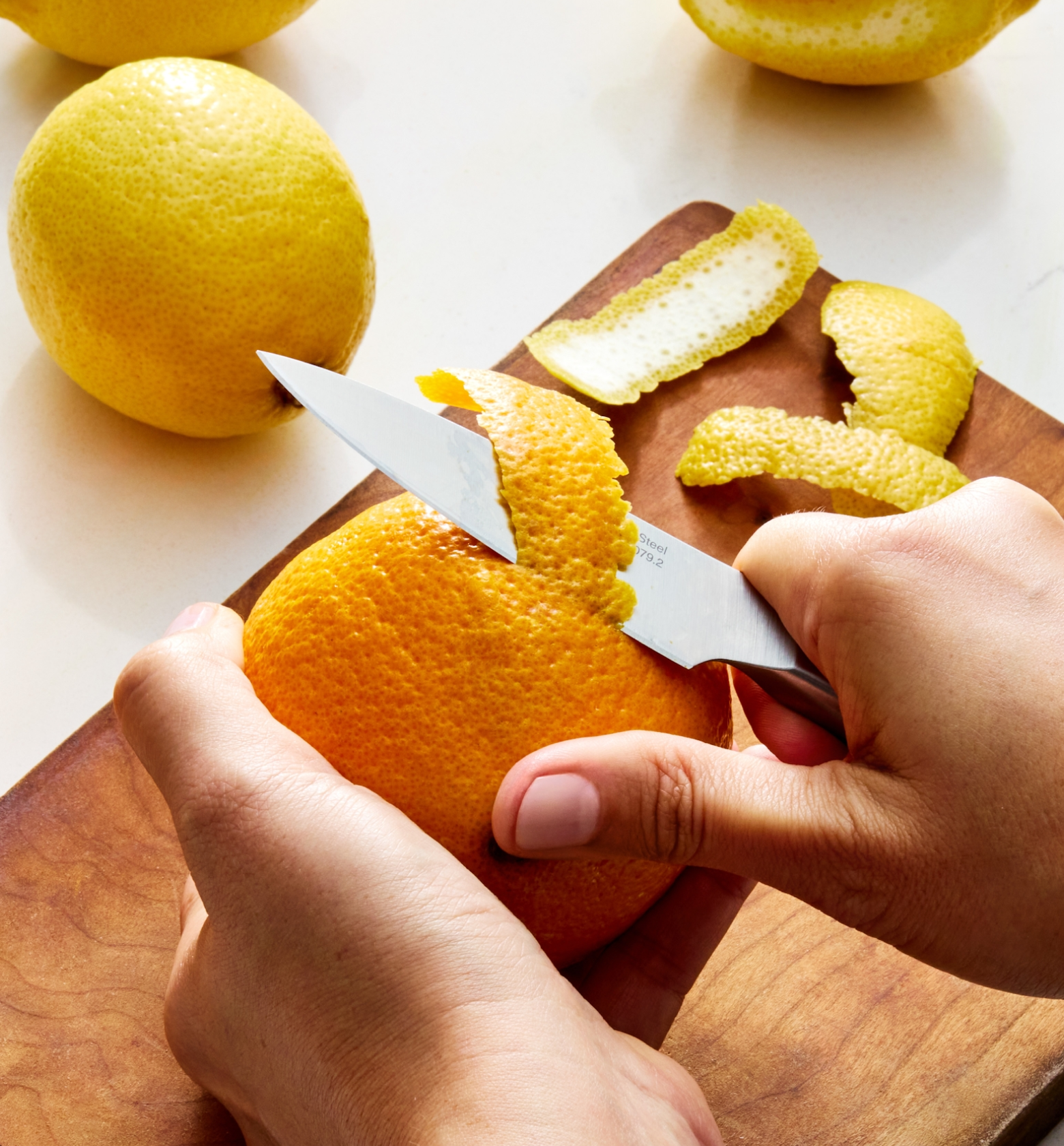 Hands peeling an orange with a paring knife on a wooden cutting board with lemon peels