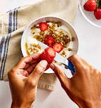 Hands slicing fresh strawberries over a yogurt granola bowl