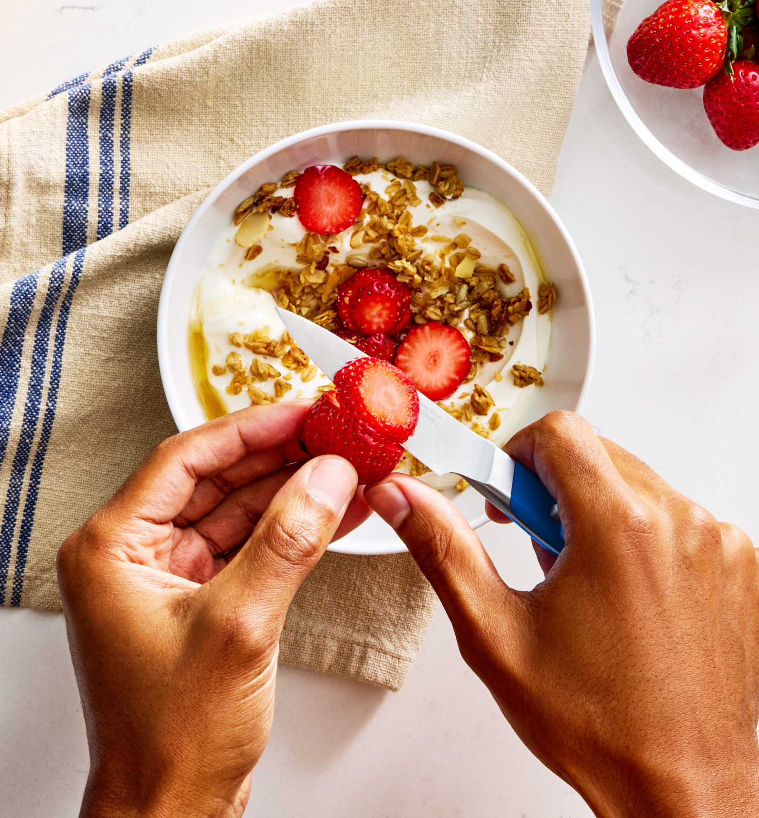 Hands slicing fresh strawberries over a yogurt granola bowl