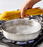 Hand adding dry spaghetti to a pan of boiling water on a gas stove