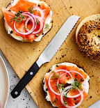 Smoked salmon bagels with cream cheese, red onion, capers, and dill on a wooden cutting board with a Misen serrated knife