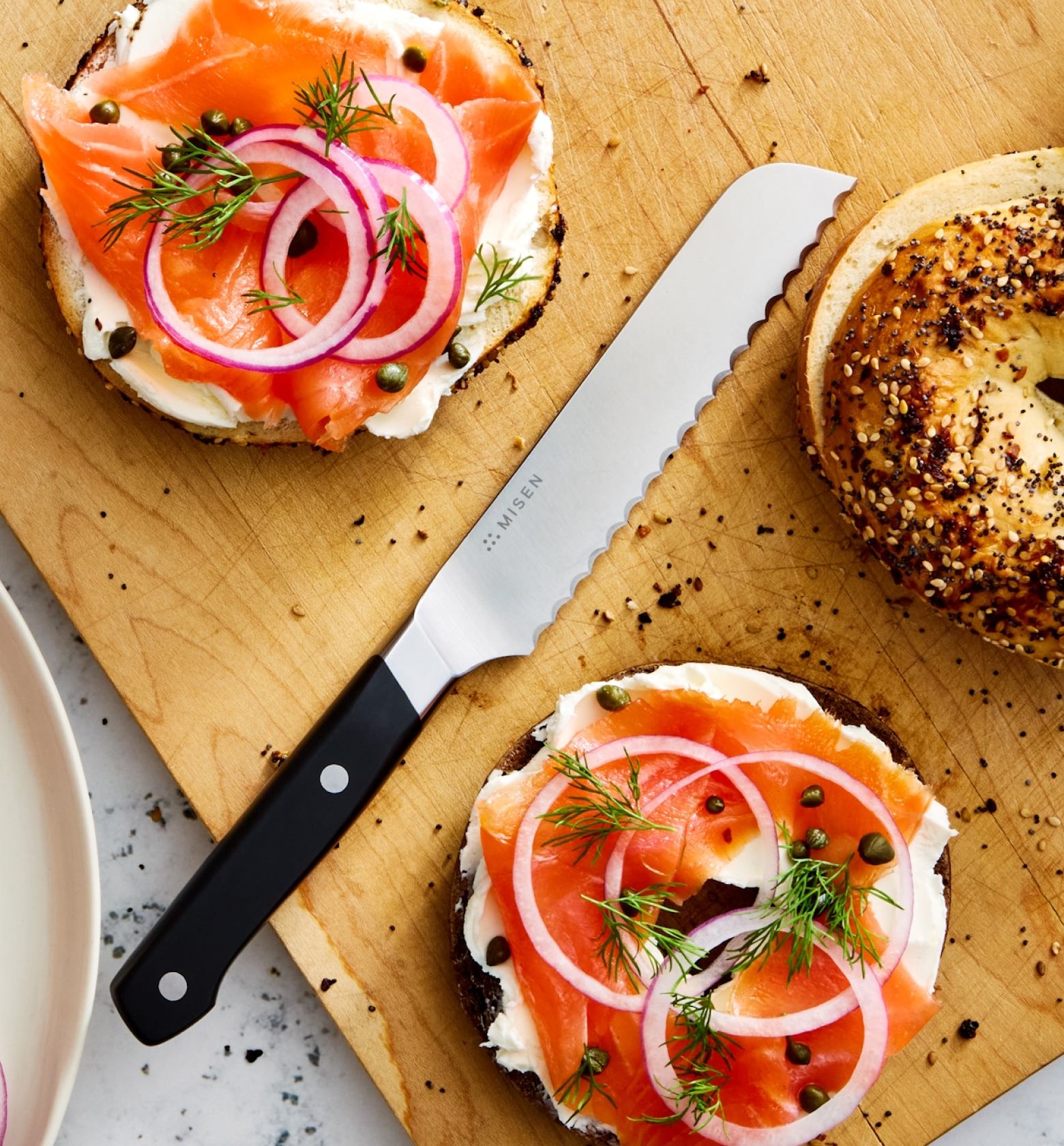 Smoked salmon bagels with cream cheese, red onion, capers, and dill on a wooden cutting board with a Misen serrated knife