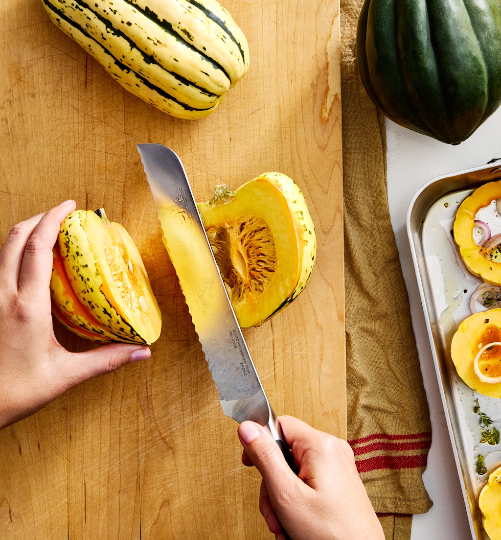 Hands cutting delicata squash on wooden cutting board with acorn squash nearby