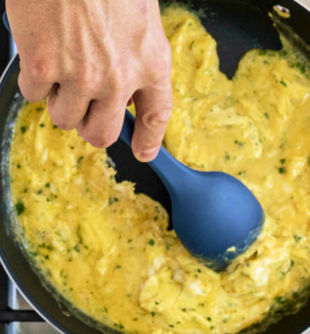 Scrambled eggs with herbs being stirred in nonstick pan with blue silicone spoon