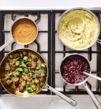 Thanksgiving side dishes on stovetop including mashed potatoes, roasted Brussels sprouts, cranberry sauce, and gravy