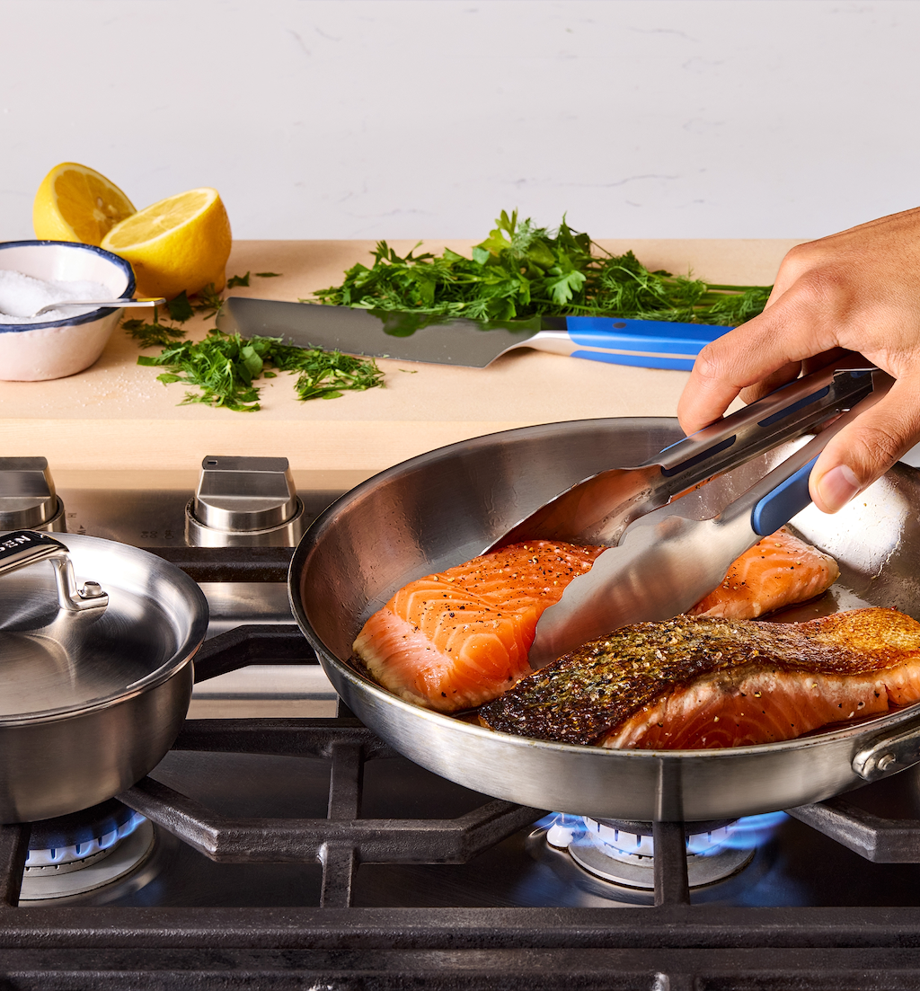 Pan-seared salmon fillets cooking in stainless steel skillet on gas stove with fresh herbs and lemon