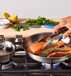 Pan-seared salmon fillets cooking in stainless steel skillet on gas stove with fresh herbs and lemon