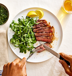 Sliced medium-rare steak with arugula salad and lemon wedges on white plate, cut with Misen steak knife