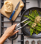 Sautéed asparagus with parmesan in a dark skillet on gas stove using Misen tongs and knife