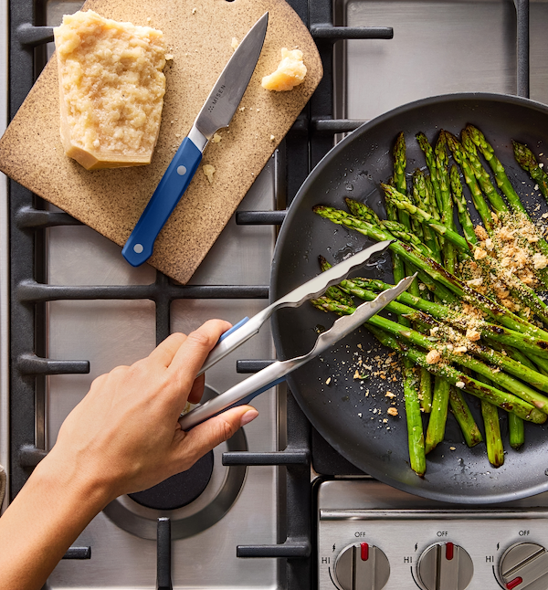 Sautéed asparagus with parmesan in a dark skillet on gas stove using Misen tongs and knife