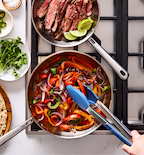 Steak fajitas with sautéed peppers and onions cooking in stainless steel pans on gas stovetop