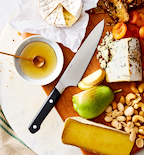 Misen chef's knife on wooden charcuterie board with brie, blue cheese, pear, nuts, honey, and dried apricots