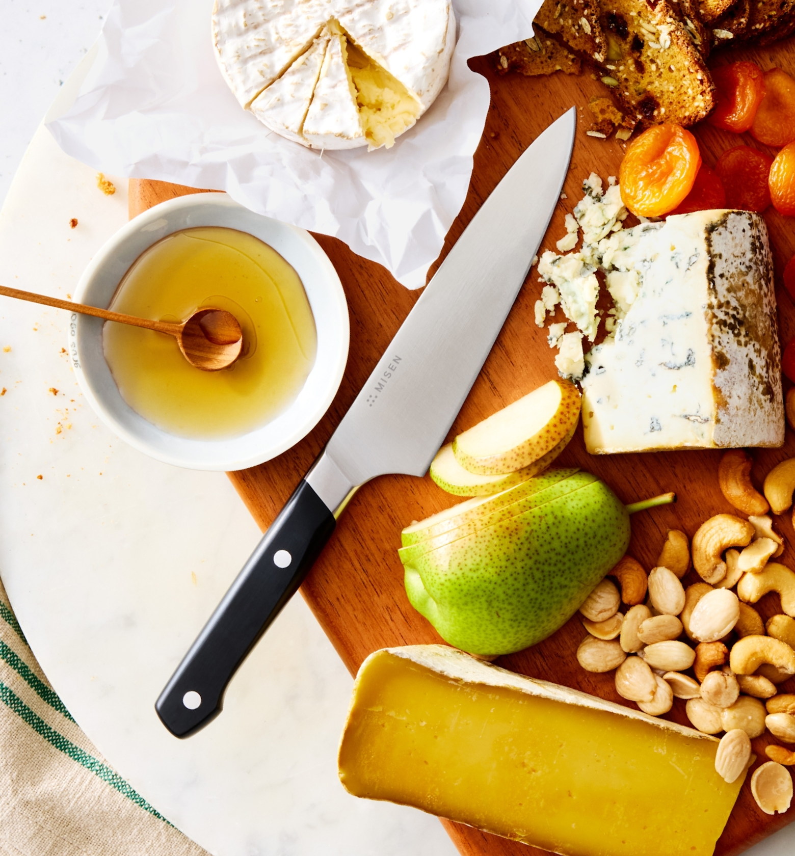 Misen chef's knife on wooden charcuterie board with brie, blue cheese, pear, nuts, honey, and dried apricots