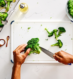 Hands cutting fresh broccoli florets on white cutting board with olive oil and salt nearby