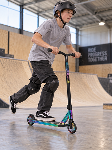 A boy wearing a helmet and knee pads rides the Legacy 1.5 Pro Ventus Complete Stunt Scooter in Rainbow Neochrome / Black by Legacy at an indoor skatepark with ramps and wooden walls.