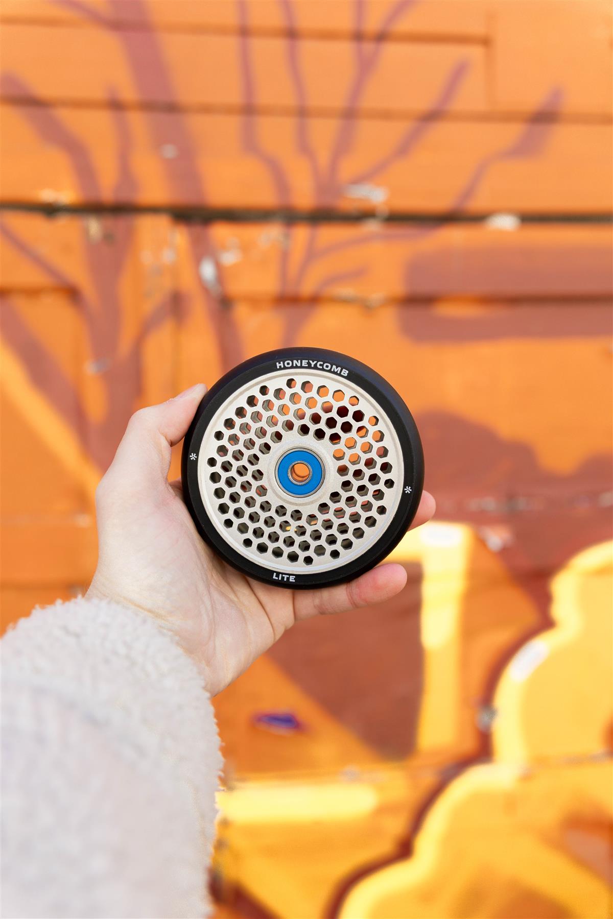 A hand holds the Union Honeycomb Lite 110mm Stunt Scooter Wheel in black and silver, featuring a perforated metal front with a honeycomb design, set against an orange mural with abstract shapes and shadows.