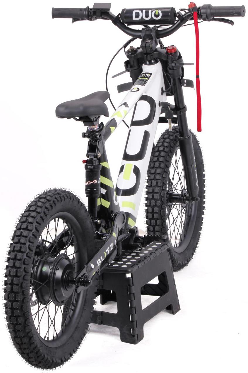 The Duo 18" Blitz Kids Electric Balance Bike in white and black, by Duo, sits on a black step stool. It features chunky tires, a padded seat, and a red strap on the handlebars, shown from the rear angle.