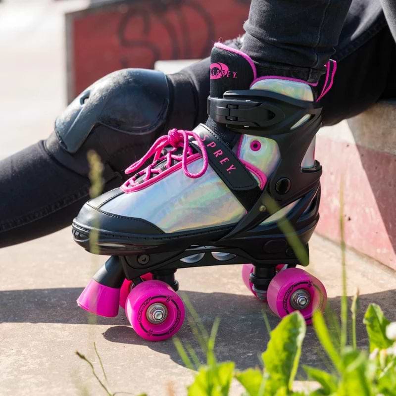 Close-up of a person wearing Osprey Surge Semi-Soft Adjustable Quad Roller Skates - Pink, featuring shiny silver and black design with bright pink wheels and laces, kneeling on concrete near a skate ramp with green plants in the foreground.