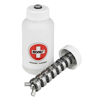 A white cylindrical container labeled with the product name "Bones Bearing Cleaning Unit," made from fluorinated high-density polyethylene, features a red cross logo associated with the Bones brand. Next to it is an opened lid and a metal rod specifically designed for the precise cleaning of skateboard bearings.