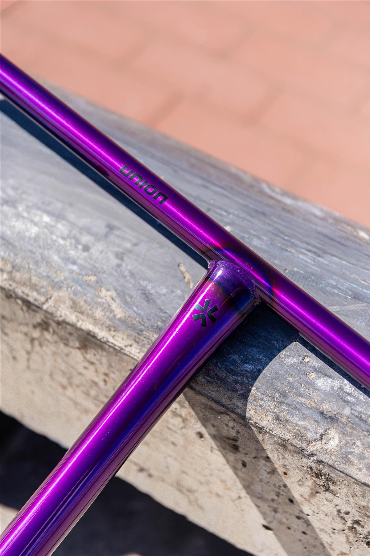 Close-up of the Union Uniq T Steel Oversized HIC Stunt Scooter Bar in polished purple (650mm x 600mm), featuring "UNION" and logo engraving, resting on a concrete ledge outdoors.