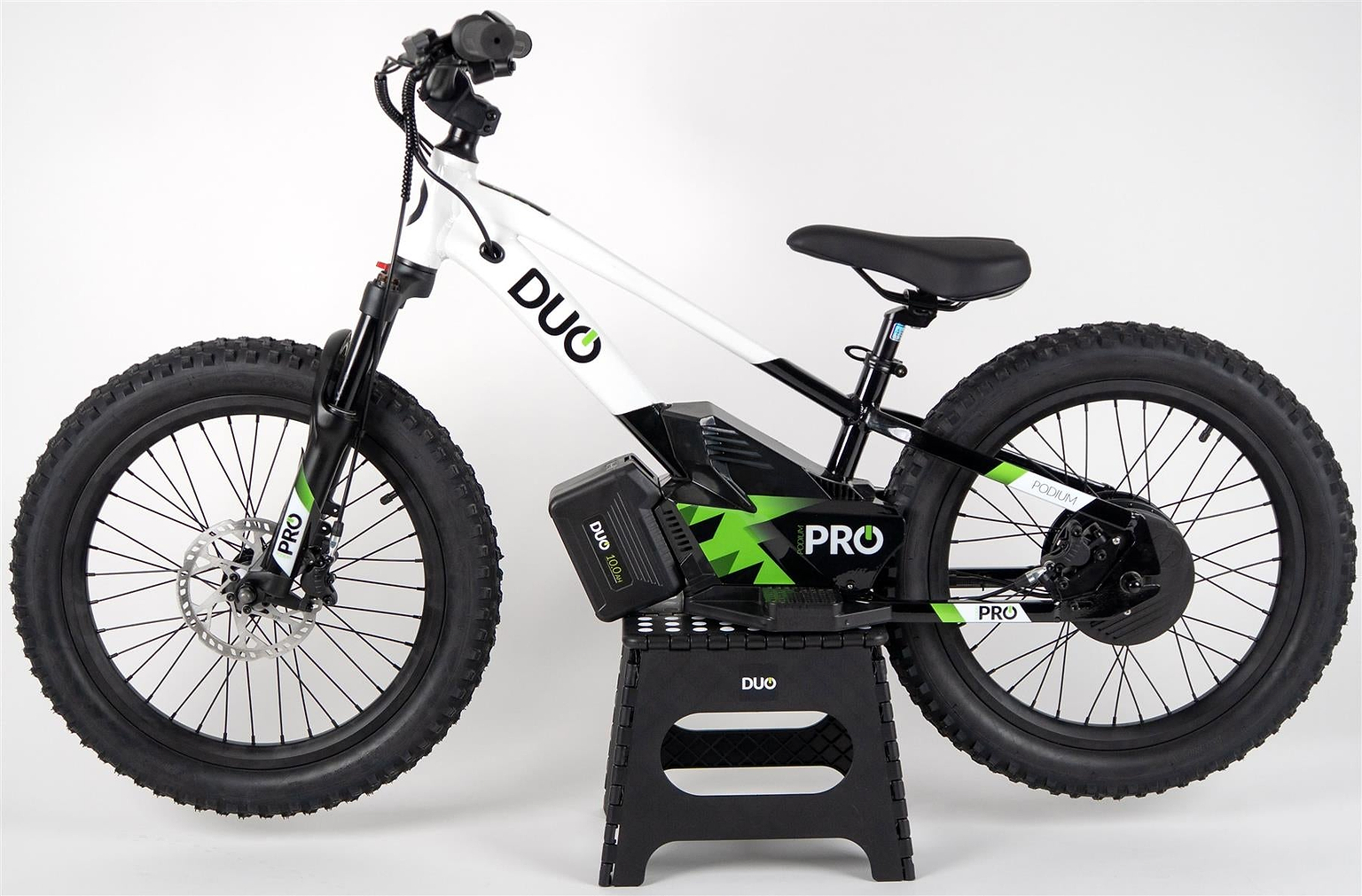 A Duo 20" Podium Pro Kids Electric Balance Bike in white and black with green accents stands on a small black stand against a plain white background. "DUO" and "PRO" are branded on the frame and battery.