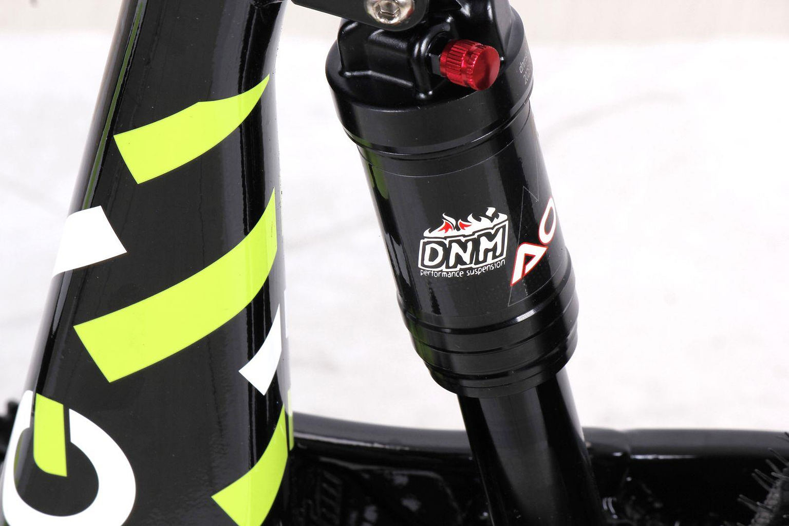 Close-up of a black DNM suspension component with the DNM logo and adjustment knob on a Duo 20" Blitz Kids Electric Balance Bike by Duo, featuring a white and black frame with green and white diagonal stripes.