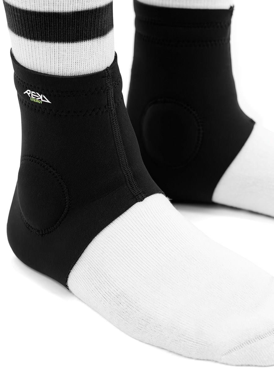 Close-up of two feet in white socks wearing REKD Energy Slim Footies Ankle Protection Pads in black, featuring zippers, circular impact padding, and a small REKD logo tag on the side.