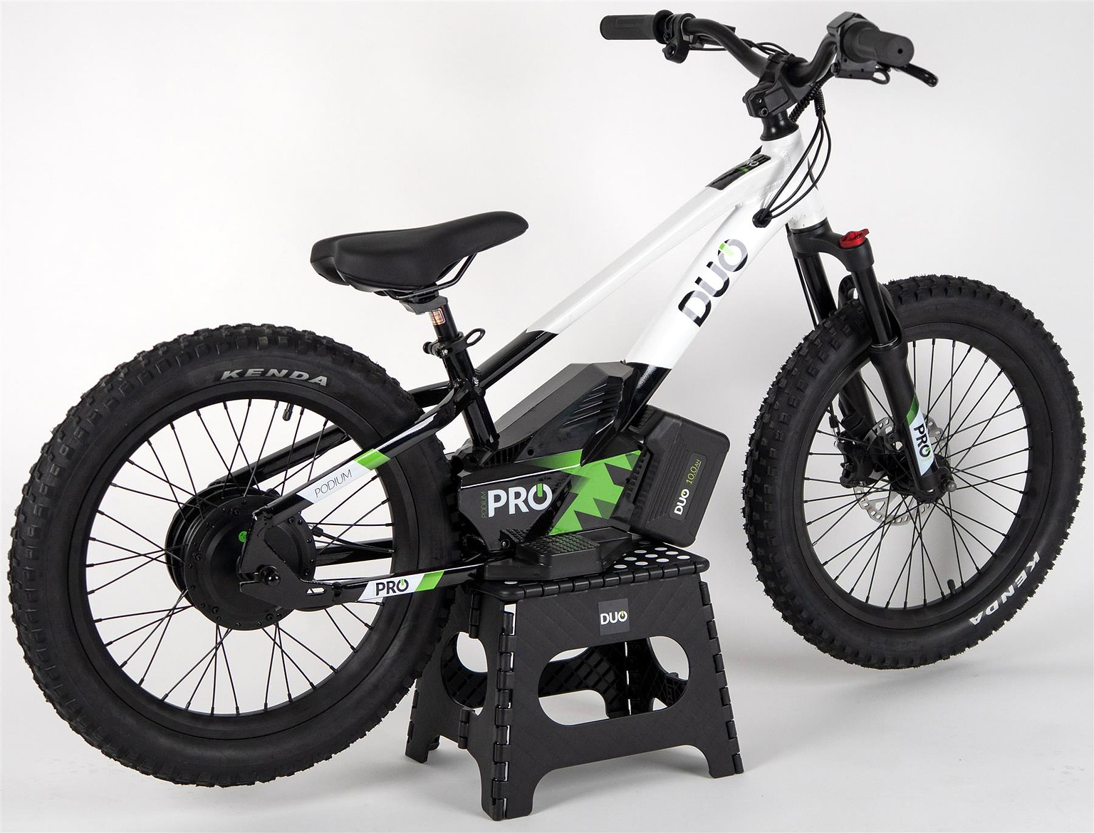 The Duo 20" Podium Pro Kids Electric Balance Bike in white and black, featuring thick tires and a battery pack for off-road use, is shown on a black stand against a plain white background.