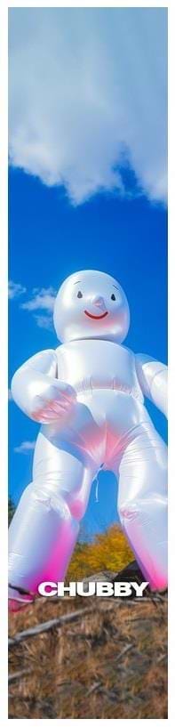 Under a bright blue sky with fluffy clouds, a tall inflatable white figure with a smiling face is positioned on the grassy area near the skatepark. At the base of this durable design, the word "Chubby" is printed, echoing the quality of Chubby's Stunt Scooter Griptape - Mellow Man.