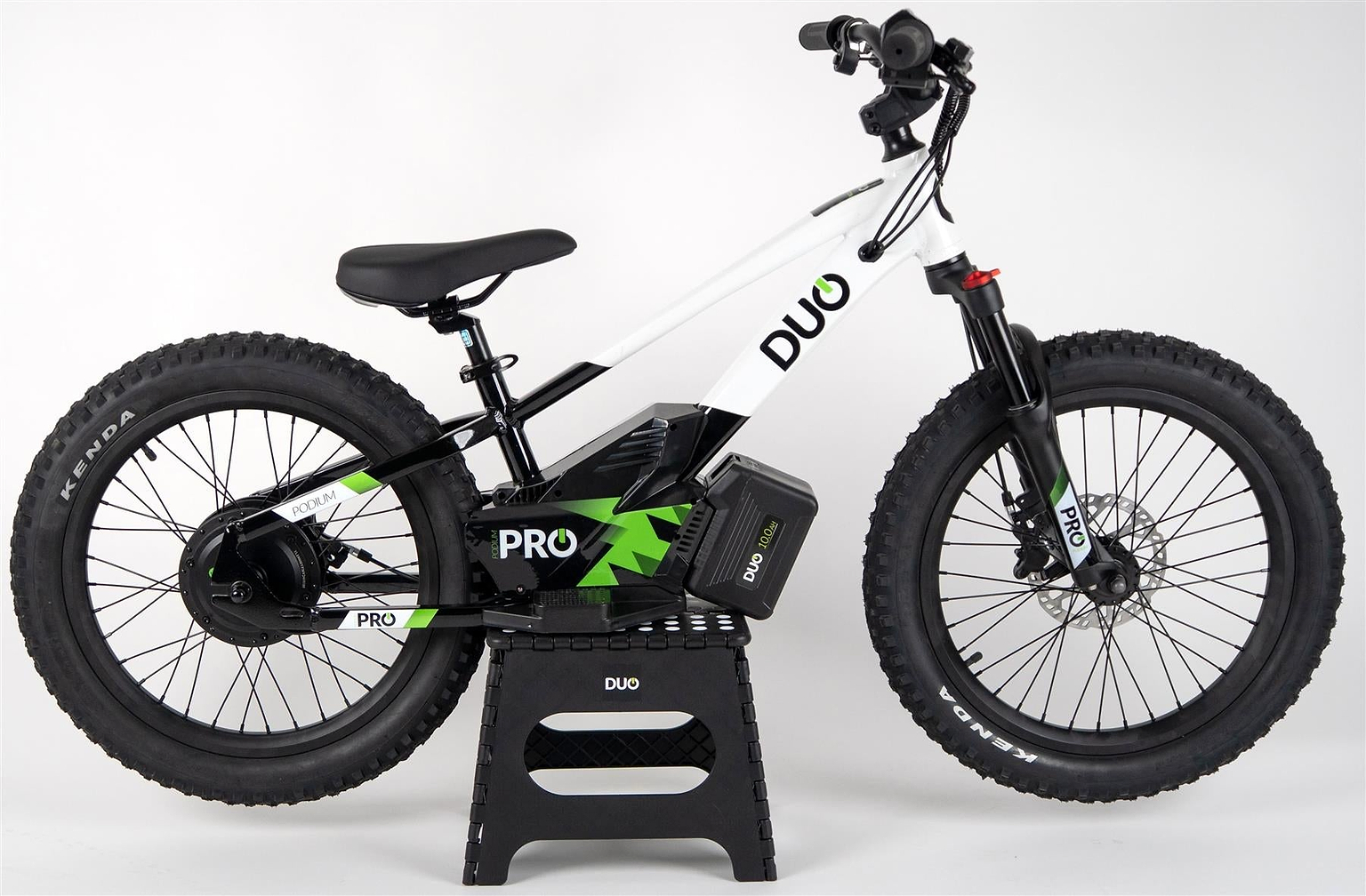 The Duo 20" Podium Pro Kids Electric Balance Bike in white and black is shown on a black stand against a plain white background, featuring a visible battery, thick tires, and sturdy frame.