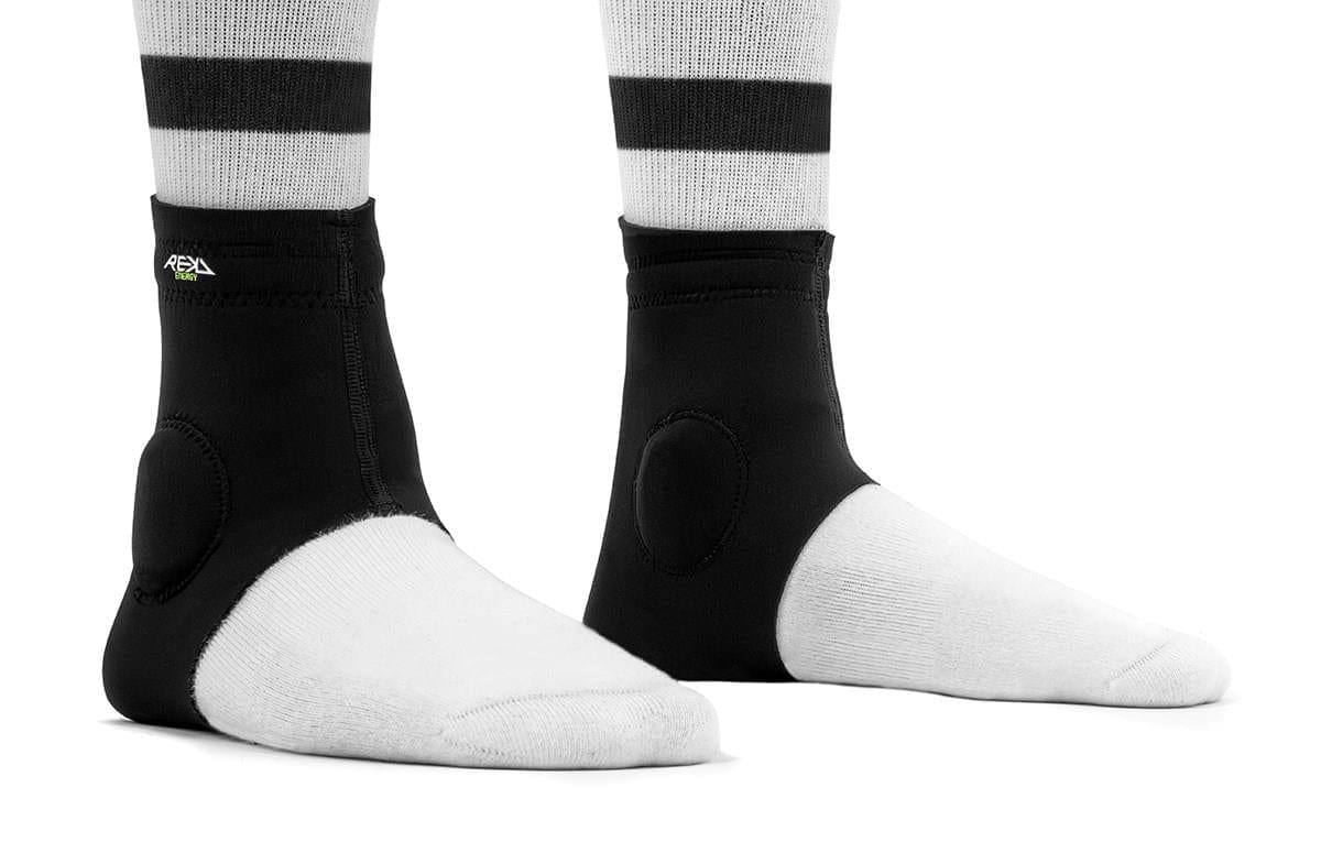 Two feet wearing REKD Energy Slim Footies Ankle Protection Pads in black, along with white socks featuring black stripes, are shown standing on a white background for ultimate ankle protection.